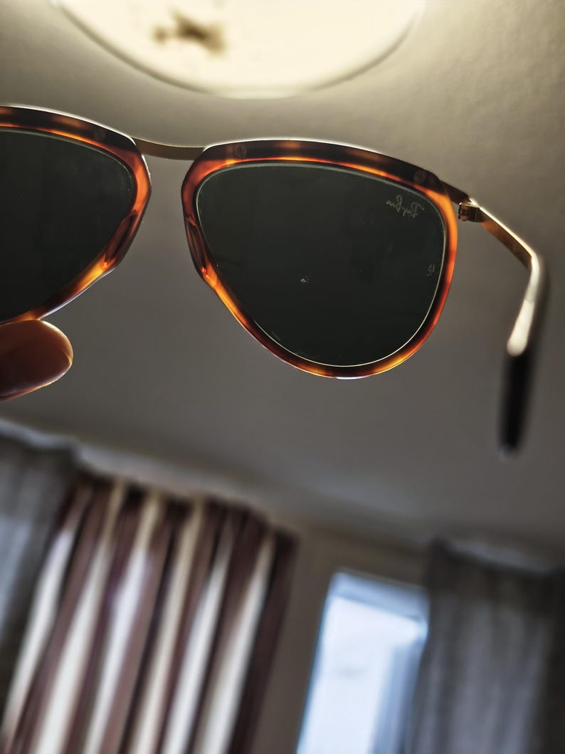 Puede incluir: Gafas de sol con montura de carey y lentes oscuras. Las gafas tienen un puente y varillas de metal dorado. El logotipo de Ray-Ban es visible en la lente.
