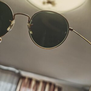 May include: Round sunglasses with dark green lenses and a thin, gold-colored metal frame. The sunglasses have a classic design with a bridge and nose pads. The lenses are set in a circular frame.