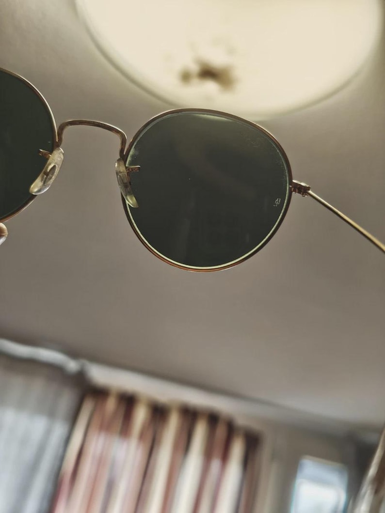 May include: Round sunglasses with dark green lenses and a thin, gold-colored metal frame. The sunglasses have a classic design with a bridge and nose pads. The lenses are set in a circular frame.