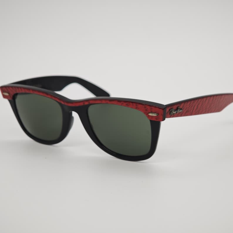 May include: Black sunglasses with red textured frames and temples. The sunglasses have dark green lenses and a classic, rectangular shape. The Ray-Ban logo is visible on the temple.
