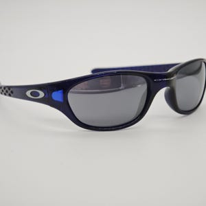 May include: Navy blue sunglasses with grey lenses. The frames have a rounded shape and a silver logo on the temples. These sunglasses are suitable for sports and outdoor activities.