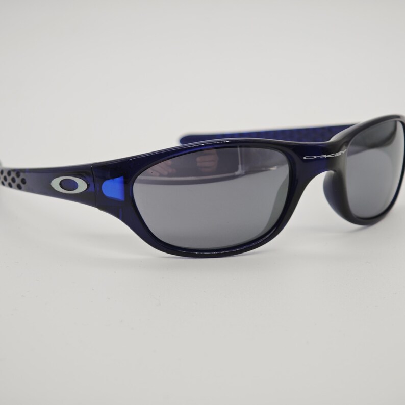 May include: Navy blue sunglasses with grey lenses. The frames have a rounded shape and a silver logo on the temples. These sunglasses are suitable for sports and outdoor activities.