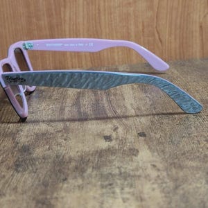 Puede incluir: Gafas de sol Ray-Ban rosas y grises con un patr&oacute;n texturizado en las patillas. Las gafas de sol tienen un estilo Wayfarer cl&aacute;sico con una montura rosa y patillas grises. Las lentes son de color marr&oacute;n.