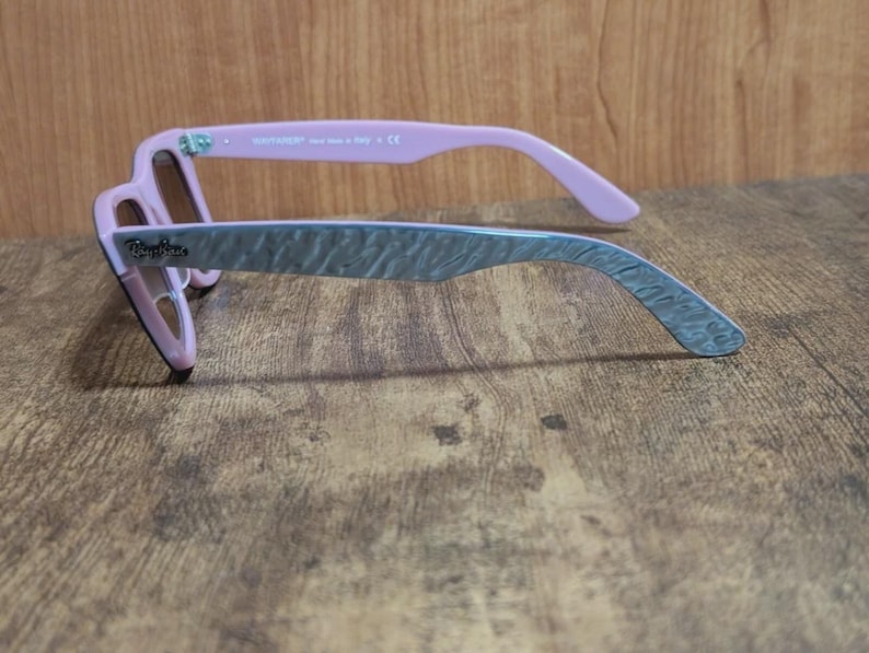 Puede incluir: Gafas de sol Ray-Ban rosas y grises con un patr&oacute;n texturizado en las patillas. Las gafas de sol tienen un estilo Wayfarer cl&aacute;sico con una montura rosa y patillas grises. Las lentes son de color marr&oacute;n.