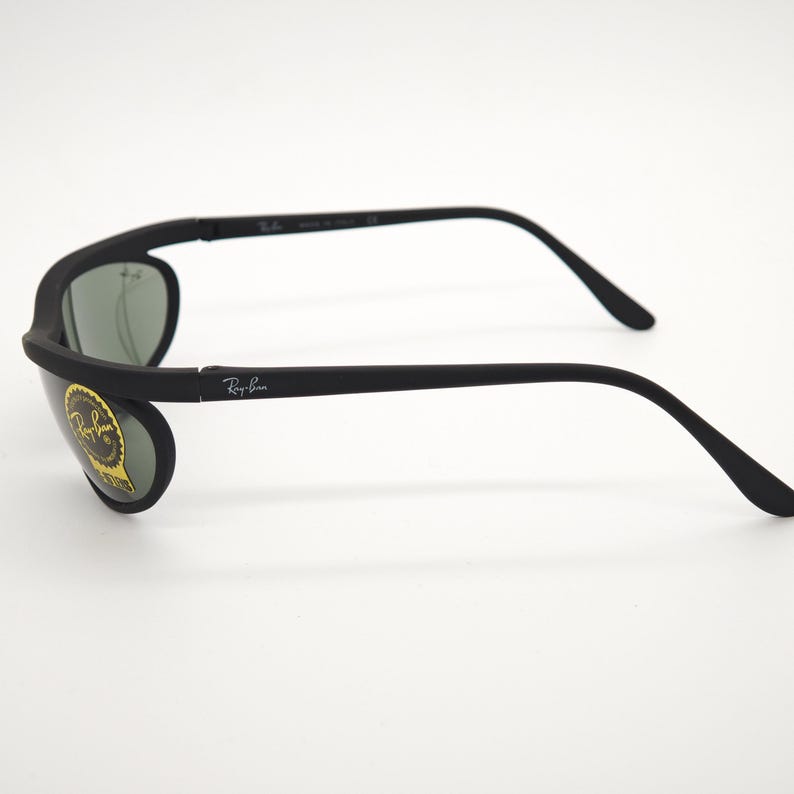 May include: Black Ray-Ban sunglasses with a matte finish. The sunglasses have a curved frame and arms, with the Ray-Ban logo on the arm. The lenses are a dark green colour. A gold and yellow sticker is on the left lens.