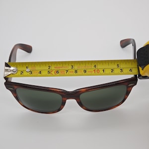 May include: Brown tortoiseshell sunglasses with green lenses. The sunglasses are measured with a tape measure, showing a width of approximately 5 inches. The frame has a classic design.