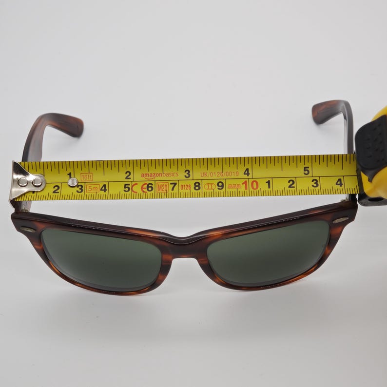 May include: Brown tortoiseshell sunglasses with green lenses. The sunglasses are measured with a tape measure, showing a width of approximately 5 inches. The frame has a classic design.