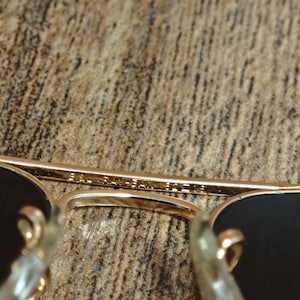 Puede incluir: Primer plano de gafas de sol con montura dorada y lentes oscuras. El puente de las gafas tiene texto grabado en el metal. Las gafas est&aacute;n sobre una superficie marr&oacute;n texturizada.
