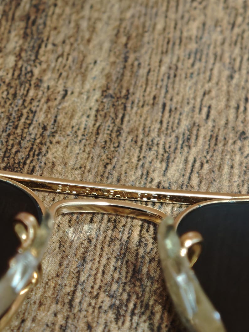 Puede incluir: Primer plano de gafas de sol con montura dorada y lentes oscuras. El puente de las gafas tiene texto grabado en el metal. Las gafas est&aacute;n sobre una superficie marr&oacute;n texturizada.