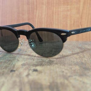 May include: Black sunglasses with a semi-rimless design. The frames are black with a gold-colored bridge and dark lenses. The arms have a textured pattern. The sunglasses are resting on a wooden surface.