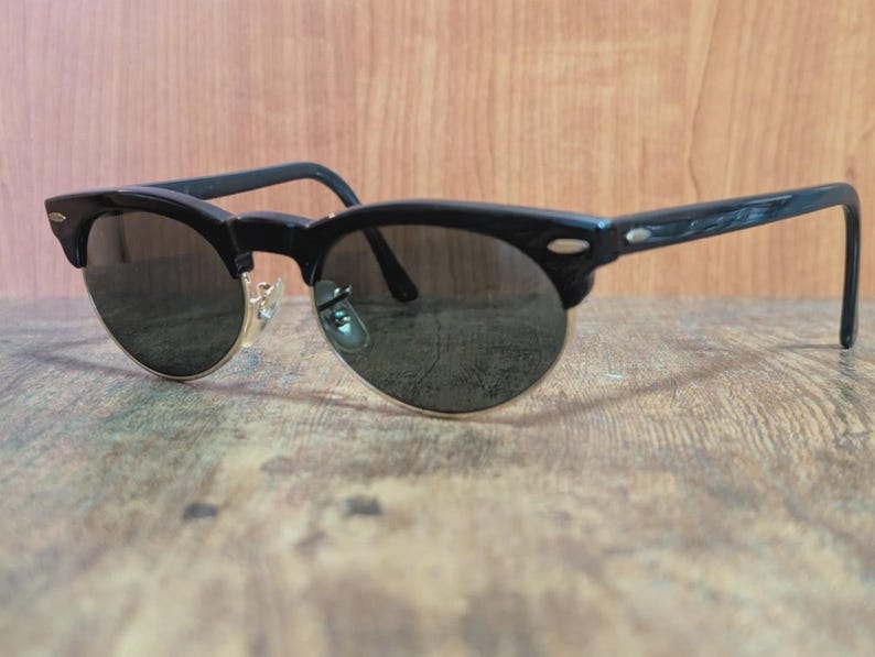 May include: Black sunglasses with a semi-rimless design. The frames are black with a gold-colored bridge and dark lenses. The arms have a textured pattern. The sunglasses are resting on a wooden surface.