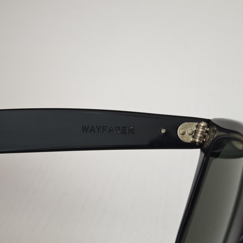 Puede incluir: Un par de gafas de sol negras con la palabra WAYFARER grabada en el interior de la patilla. La montura incluye una bisagra met&aacute;lica de tono plateado con dos remaches visibles que conectan la patilla con la parte frontal de la montura.