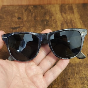 Puede incluir: Gafas de sol con montura oscura y veteada y lentes negras. Las gafas de sol tienen una forma rectangular cl&aacute;sica. La montura tiene una mezcla de tonos azul oscuro y gris, lo que le da una apariencia &uacute;nica y texturizada. Las lentes son oscuras, proporcionando sombra y protecci&oacute;n solar.