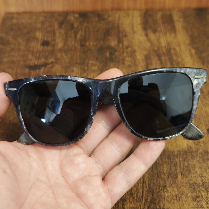 Puede incluir: Gafas de sol con montura oscura y veteada y lentes negras. Las gafas de sol tienen una forma rectangular cl&aacute;sica. La montura tiene una mezcla de tonos azul oscuro y gris, lo que le da una apariencia &uacute;nica y texturizada. Las lentes son oscuras, proporcionando sombra y protecci&oacute;n solar.