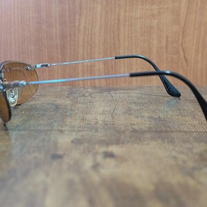 May include: Sunglasses with brown tinted lenses and a silver frame. The temples are black and curve down towards the ears. The sunglasses are resting on a wooden surface.