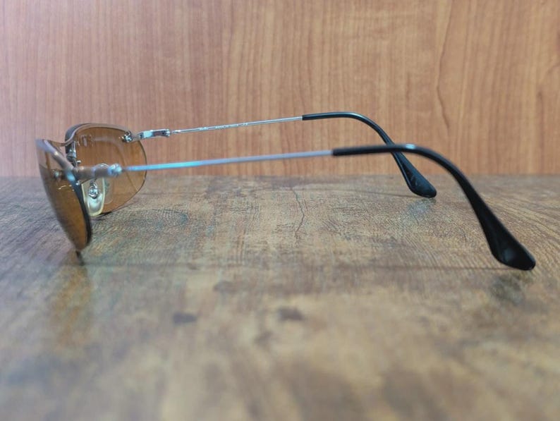 May include: Sunglasses with brown tinted lenses and a silver frame. The temples are black and curve down towards the ears. The sunglasses are resting on a wooden surface.