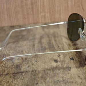 May include: Silver-framed aviator sunglasses with dark lenses. The sunglasses are positioned on a wooden surface, showcasing the sleek design of the frames and the shape of the lenses. The arms of the glasses extend outwards.