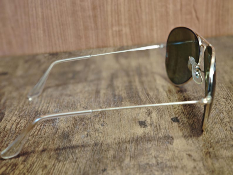 May include: Silver-framed aviator sunglasses with dark lenses. The sunglasses are positioned on a wooden surface, showcasing the sleek design of the frames and the shape of the lenses. The arms of the glasses extend outwards.