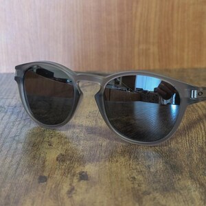 May include: Gray sunglasses with dark lenses. The sunglasses have a rounded frame and a sleek, modern design. The lenses reflect the surrounding environment. The sunglasses are resting on a wooden surface.