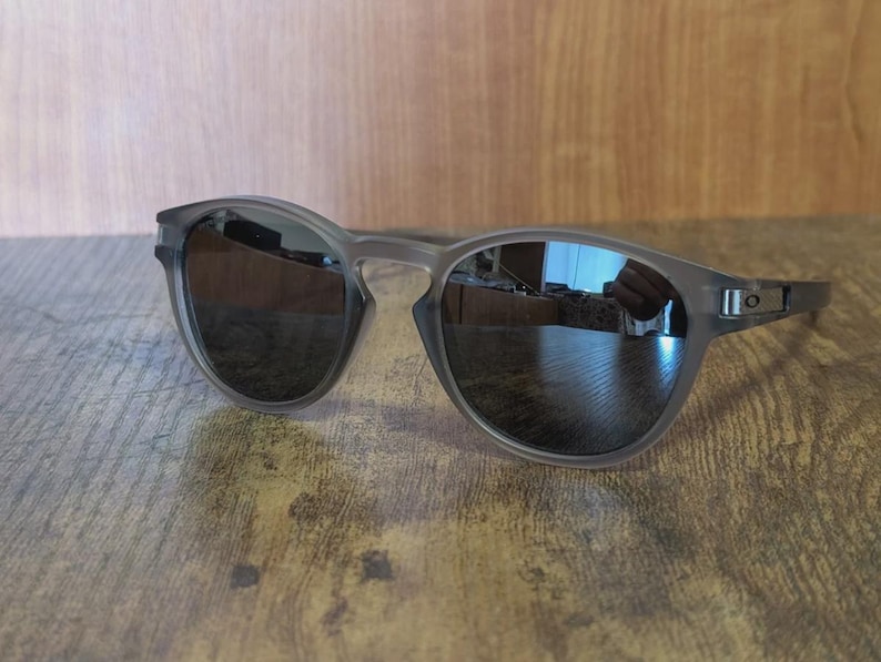 May include: Gray sunglasses with dark lenses. The sunglasses have a rounded frame and a sleek, modern design. The lenses reflect the surrounding environment. The sunglasses are resting on a wooden surface.