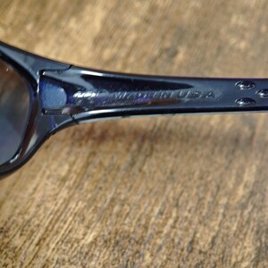 May include: Close-up of dark blue sunglasses. The temple arm is visible, with the text "MADE IN USA" printed on it. The sunglasses are set against a wooden background.