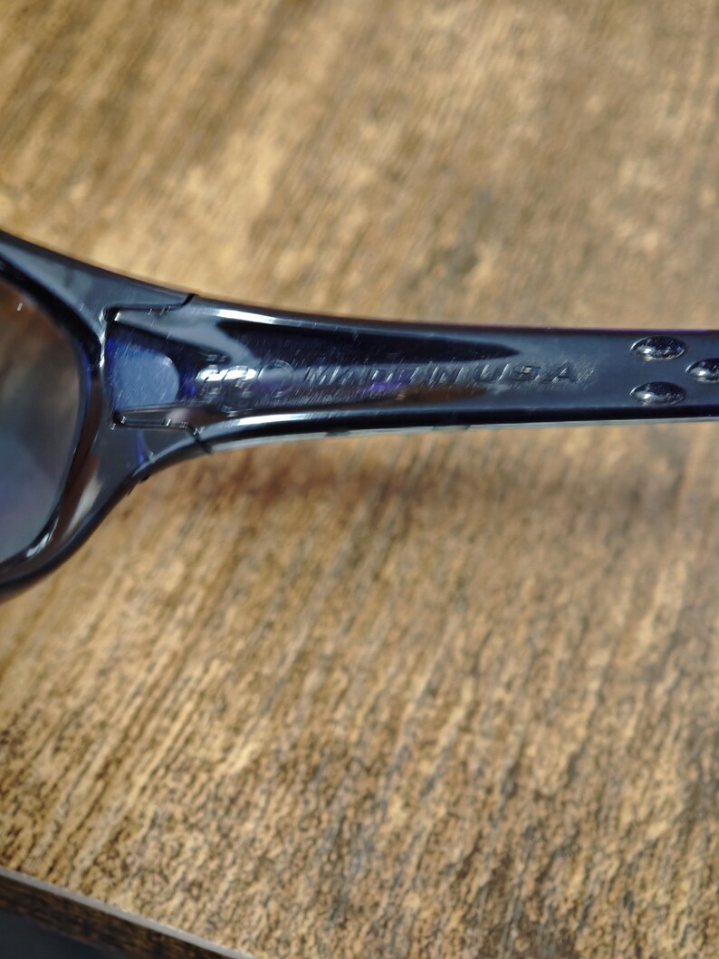 May include: Close-up of dark blue sunglasses. The temple arm is visible, with the text "MADE IN USA" printed on it. The sunglasses are set against a wooden background.