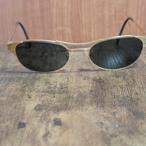 May include: Gold-toned sunglasses with dark lenses. The sunglasses have a unique, curved frame design and black temples. The Ray-Ban logo is visible on the lens. The sunglasses are set against a wooden background.