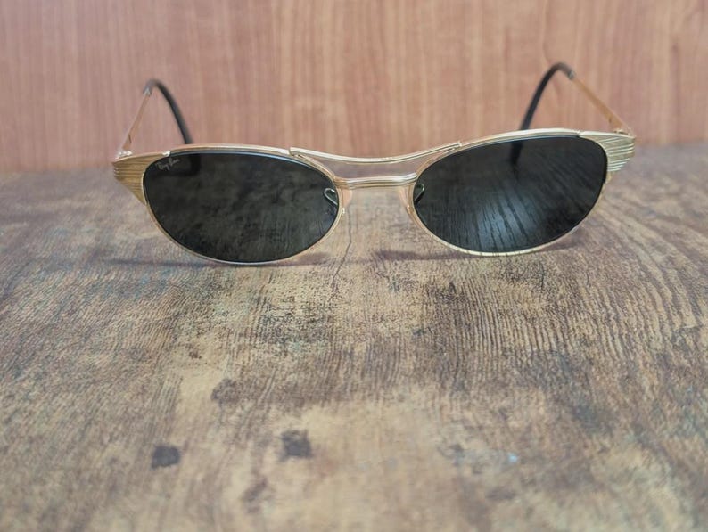 May include: Gold-toned sunglasses with dark lenses. The sunglasses have a unique, curved frame design and black temples. The Ray-Ban logo is visible on the lens. The sunglasses are set against a wooden background.