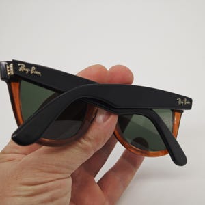 May include: Black and orange Ray-Ban sunglasses with green lenses. The sunglasses have a classic design with the Ray-Ban logo on the temples. The frames are black on top and orange on the bottom.
