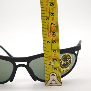 May include: Black Ray-Ban sunglasses with green lenses. A yellow tape measure shows the lenses are approximately 5 cm in height. The sunglasses have a sticker that says "Ray-Ban".