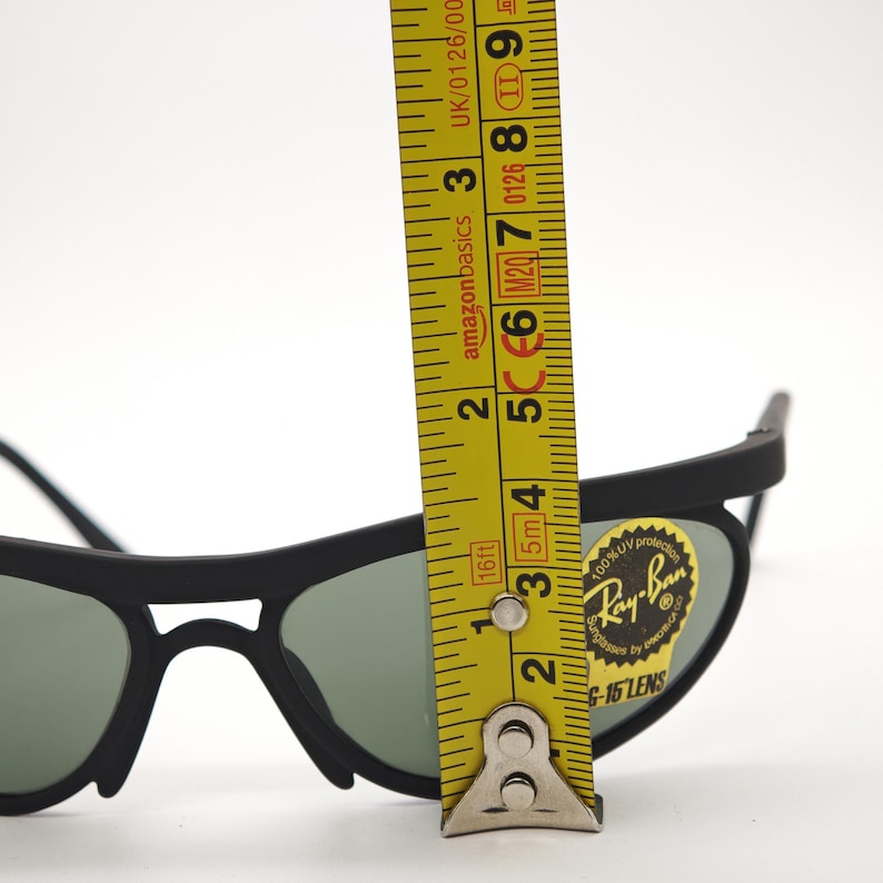 May include: Black Ray-Ban sunglasses with green lenses. A yellow tape measure shows the lenses are approximately 5 cm in height. The sunglasses have a sticker that says "Ray-Ban".