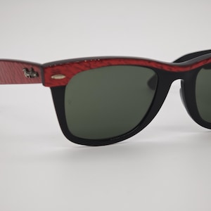 May include: Black sunglasses with a red patterned top frame and dark green lenses. The sunglasses have a classic design with a black frame and a small silver emblem on the side.