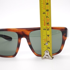 May include: Tortoiseshell sunglasses with green lenses. The frame is measured with a yellow tape measure. The sunglasses are approximately 2 inches in height.