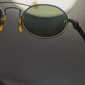 May include: Close-up of vintage sunglasses with round, green-tinted lenses. The frames are a dark metal with tortoise-shell accents on the temples. The lenses have a small logo on the right lens.
