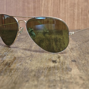 May include: Aviator sunglasses with a gold-colored frame and green-tinted lenses. The sunglasses are resting on a wooden surface. The lenses reflect the surrounding environment.