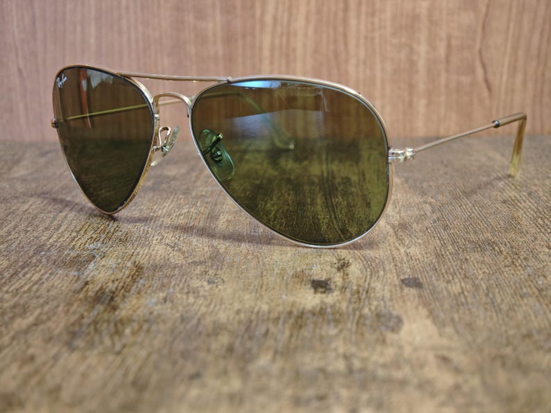 May include: Aviator sunglasses with a gold-colored frame and green-tinted lenses. The sunglasses are resting on a wooden surface. The lenses reflect the surrounding environment.