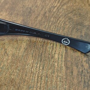K&ouml;nnte beinhalten: Nahaufnahme des B&uuml;gels einer schwarzen Sonnenbrille. Der Text "MADE IN U.S.A." ist sichtbar. Ein wei&szlig;es Emblem befindet sich am B&uuml;gel. Die Sonnenbrille liegt auf einer Holzoberfl&auml;che.