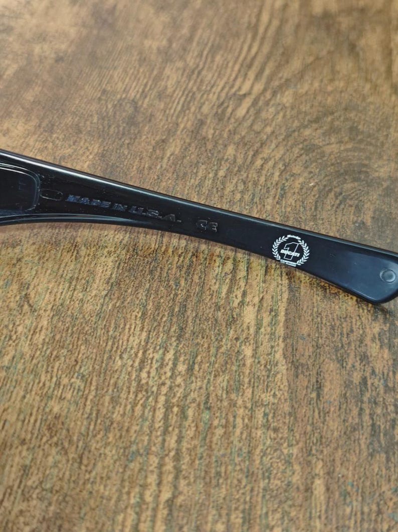 K&ouml;nnte beinhalten: Nahaufnahme des B&uuml;gels einer schwarzen Sonnenbrille. Der Text "MADE IN U.S.A." ist sichtbar. Ein wei&szlig;es Emblem befindet sich am B&uuml;gel. Die Sonnenbrille liegt auf einer Holzoberfl&auml;che.