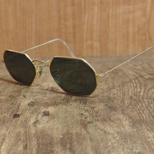 May include: Gold-rimmed sunglasses with dark lenses. The glasses have a unique octagonal shape and thin, gold-colored arms. The lenses are a dark shade, providing contrast against the gold frames. The sunglasses are resting on a wooden surface.