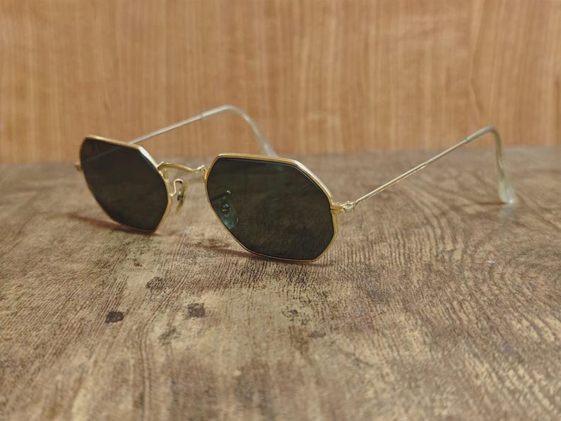 May include: Gold-rimmed sunglasses with dark lenses. The glasses have a unique octagonal shape and thin, gold-colored arms. The lenses are a dark shade, providing contrast against the gold frames. The sunglasses are resting on a wooden surface.