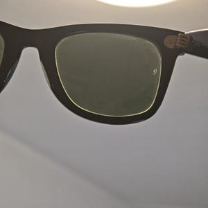 May include: Close-up of black sunglasses with dark green lenses. The frames are thick and rectangular, with a subtle gold trim around the lenses. The hinges are visible, and the overall design is classic and timeless.