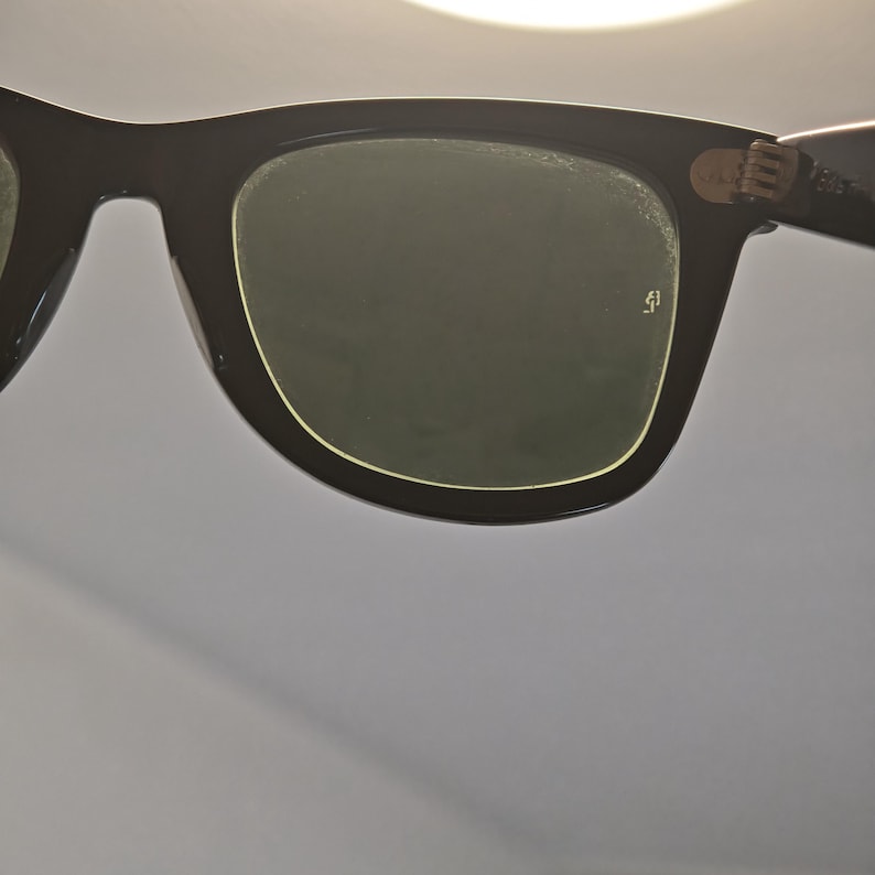 May include: Close-up of black sunglasses with dark green lenses. The frames are thick and rectangular, with a subtle gold trim around the lenses. The hinges are visible, and the overall design is classic and timeless.