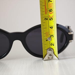 Puede incluir: Gafas de sol ovaladas negras con lentes oscuras y detalles met&aacute;licos plateados en las patillas. Una cinta m&eacute;trica amarilla est&aacute; colocada verticalmente sobre la lente derecha, mostrando una altura de marco de unos 3,8 cm.