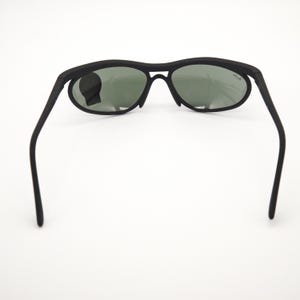 May include: Black sunglasses with oval lenses and a matte finish. The sunglasses have a sleek, modern design with a bridge and arms. The lenses are a dark green colour. The Ray-Ban logo is visible on the right lens.