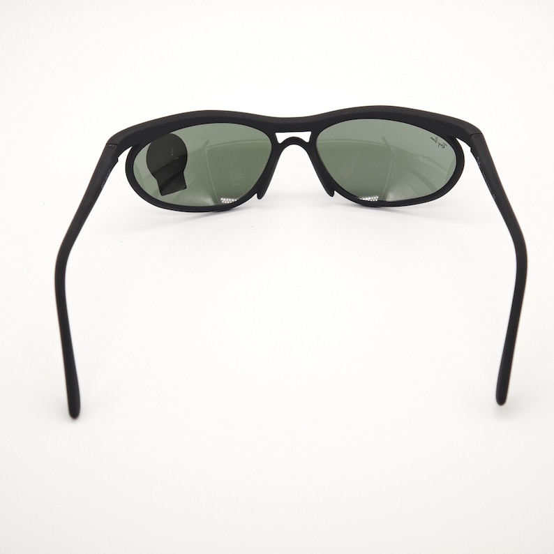 May include: Black sunglasses with oval lenses and a matte finish. The sunglasses have a sleek, modern design with a bridge and arms. The lenses are a dark green colour. The Ray-Ban logo is visible on the right lens.
