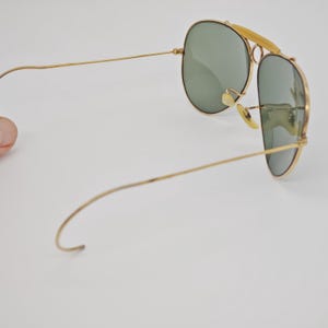 May include: Gold-framed aviator sunglasses with green lenses. The sunglasses have a double bridge and a curved brow bar. The temples are thin and extend to the ear pieces. The lenses are oval-shaped.