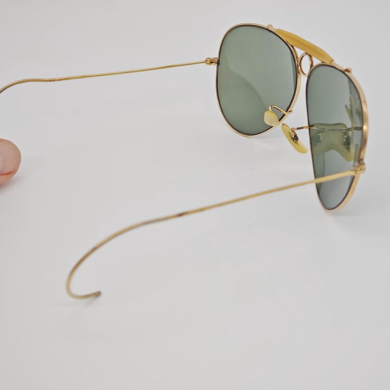 May include: Gold-framed aviator sunglasses with green lenses. The sunglasses have a double bridge and a curved brow bar. The temples are thin and extend to the ear pieces. The lenses are oval-shaped.