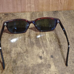 May include: Sunglasses with a dark tortoiseshell frame and dark lenses. The rectangular frames have a classic design, with black arms extending from the frame. The sunglasses are set against a textured, brown surface.