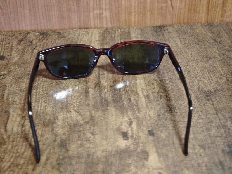 May include: Sunglasses with a dark tortoiseshell frame and dark lenses. The rectangular frames have a classic design, with black arms extending from the frame. The sunglasses are set against a textured, brown surface.