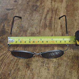 May include: A pair of oval-shaped sunglasses with dark lenses and thin, dark metal frames. The glasses are measured with a tape measure, showing a length of approximately 5 inches.